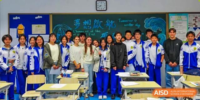 A diverse group of students in both casual clothes and blue school uniforms poses for a friendly group photo in front of a chalkboard at Beijing LuHe International Academy.