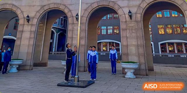 A group of students in matching blue tracksuits stands at attention during a formal flag-raising ceremony held in a stone courtyard at Beijing LuHe International Academy.