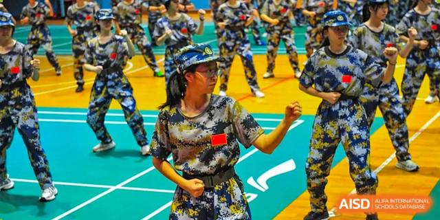 A focused female student in a blue and yellow camouflage uniform practices martial arts stances during a rigorous physical training session held indoors at Beijing LuHe International Academy.