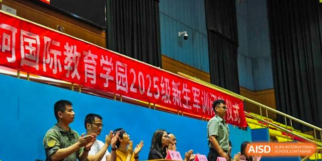 Faculty members and guests stand and clap during a formal opening ceremony featuring a large red banner for the 2025 student military training program at Beijing LuHe International Academy.