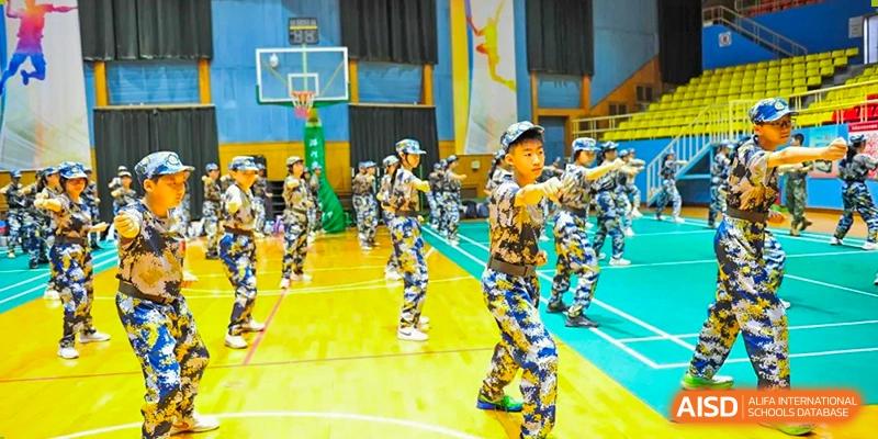 A large group of students in full camouflage attire practices martial arts punch techniques in a spacious gymnasium with a basketball hoop in the background at Beijing LuHe International Academy.