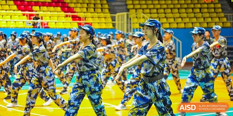 Two female students in camouflage gear lead a group through synchronized movements and drills on a blue indoor court during a physical education session at Beijing LuHe International Academy.
