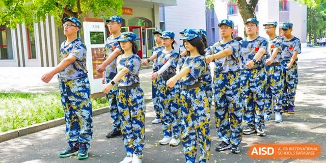 A squad of students in blue camouflage caps and uniforms practices marching in unison during an outdoor training session on a paved area of the campus at Beijing LuHe International Academy.