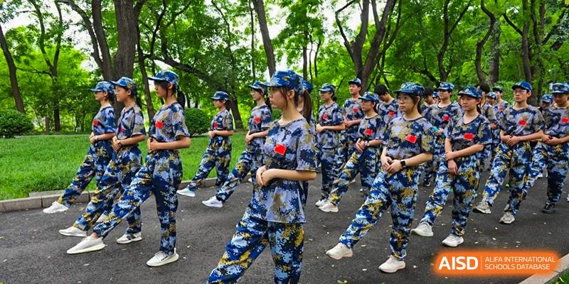 Students in camouflage uniforms practice marching in formation along a tree-lined campus path during a sunny outdoor training session held at Beijing LuHe International Academy.