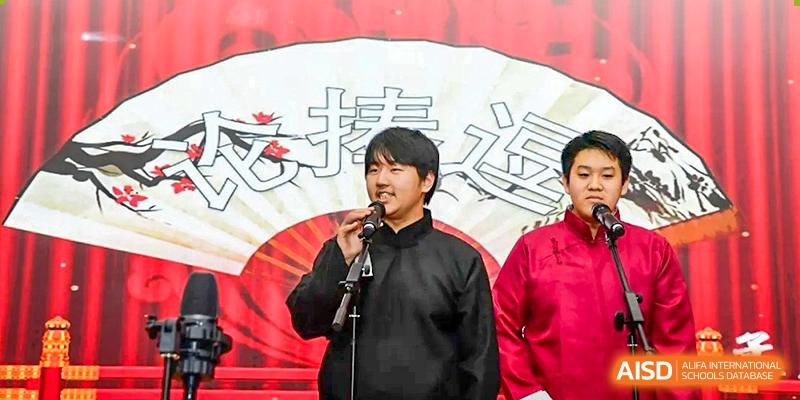 Two male students in traditional dark robes perform a comedic dialogue or speech on a stage in front of a large decorative fan backdrop at Beijing LuHe International Academy.