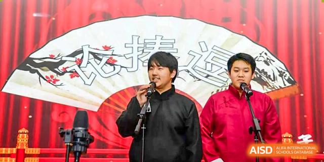 Two male students in traditional dark robes perform a comedic dialogue or speech on a stage in front of a large decorative fan backdrop at Beijing LuHe International Academy.