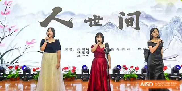 Two female vocalists in elegant dresses perform a duet on a stage featuring a backdrop with traditional Chinese calligraphy during a school talent show at Beijing LuHe International Academy