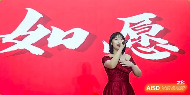 A female student in a red dress performs a vocal solo with passion on a stage featuring a bold red backdrop with traditional calligraphy at Beijing LuHe International Academy.