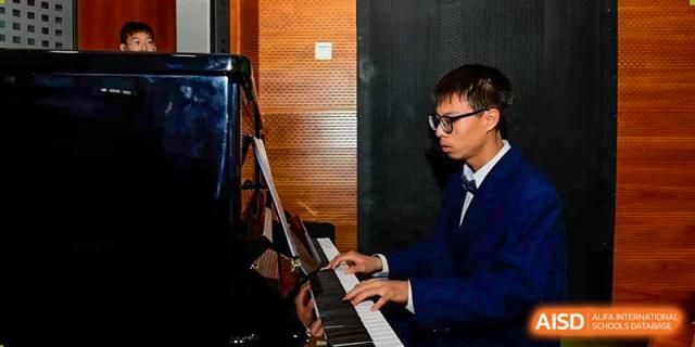 A male student wearing a formal blue suit and glasses focuses intently while playing a classical piece on a black grand piano during a recital at Beijing LuHe International Academy.