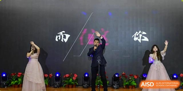 A male student dressed in a formal black suit delivers a speech or song with an expressive hand gesture on a professional stage at Beijing LuHe International Academy.