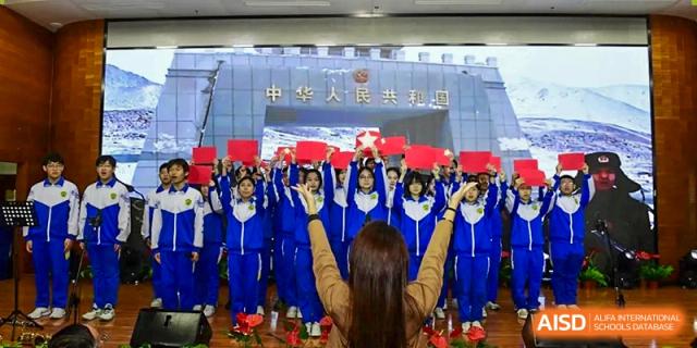 A large group of students in blue and white uniforms stands on stage with their hands raised while holding red banners during a ceremony at Beijing LuHe International Academy.