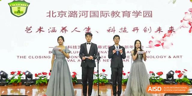 Four student presenters in formal attire stand on a stage with microphones to host a ceremony in front of a large white commemorative banner at Beijing LuHe International Academy.