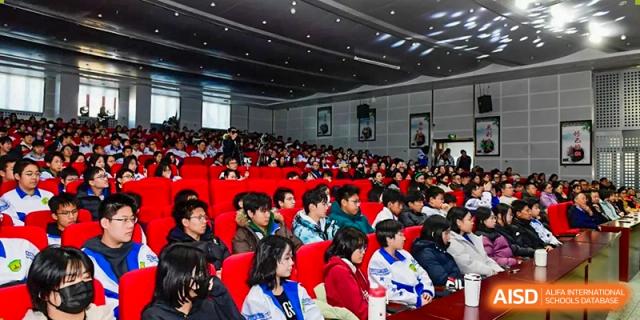 A large audience of students in various winter jackets and school uniforms sits in red auditorium seats, watching a performance at Beijing LuHe International Academy.