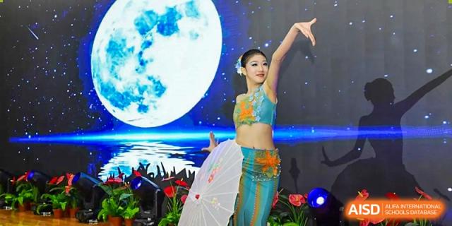 A female student in a traditional blue and green costume performs a classical dance with a white umbrella against a backdrop of a full moon at Beijing LuHe International Academy.