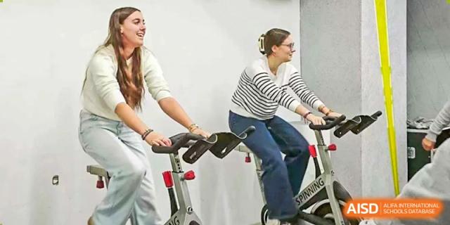 Two female students smile brightly while engaging in a high-energy cardiovascular workout on stationary spinning bikes inside a well-equipped fitness center at Beijing LuHe International Academy.