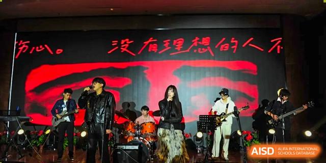 A student rock band featuring a lead singer, drummer, and guitarist performs a high-energy set in front of a red and black graphic backdrop at Beijing LuHe International Academy.