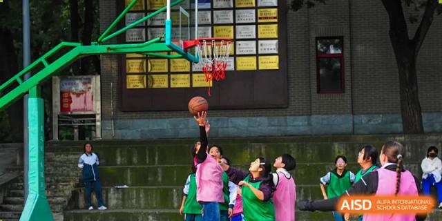 A student in a pink jersey jumps to take a shot at the basketball hoop while other players watch during a competitive outdoor sports match on the court at Beijing LuHe International Academy.