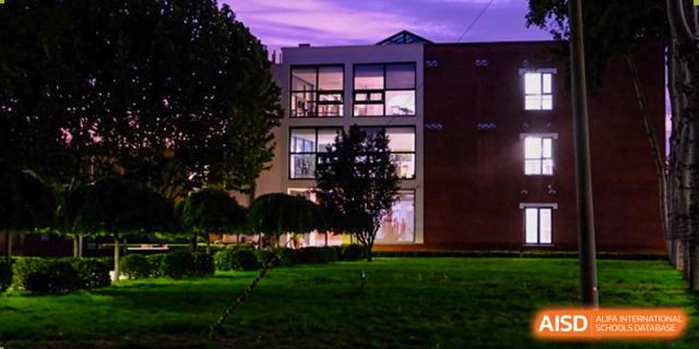 The modern brick exterior of the school building is illuminated at night, showing large glass windows and a well-maintained green lawn in the foreground at Beijing Tongzhou Shuren Private School.