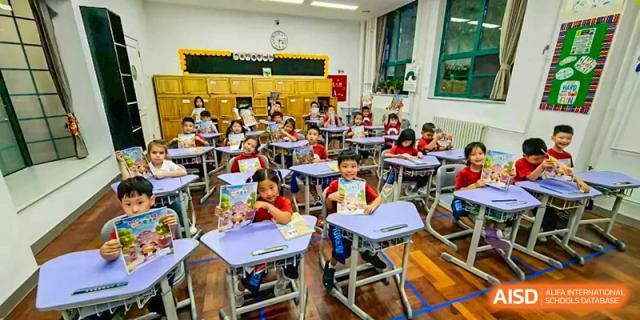 Students sit at their blue desks in a vibrant classroom, holding up colorful workbooks and engaging actively in their primary school lesson at Beijing Tongzhou Shuren Private School.