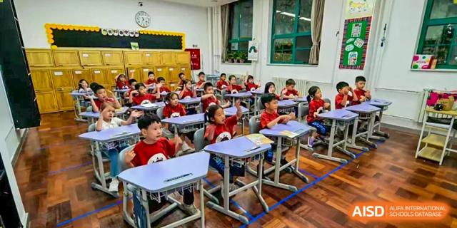 A classroom of young students in red shirts sits attentively at their desks, listening to a lesson in a bright room with educational posters at Beijing Tongzhou Shuren Private School.