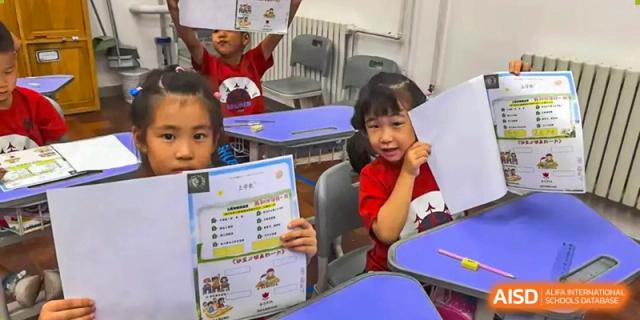 Two young students enthusiastically hold up their completed classroom assignments and drawings, showcasing their creative progress at Beijing Tongzhou Shuren Private School.