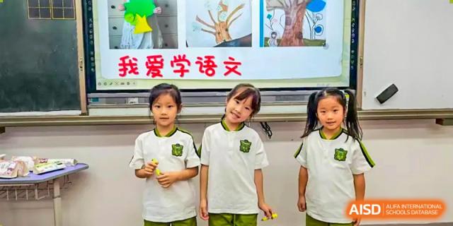 Three young female students in matching white and green uniforms stand proudly in front of a whiteboard displaying their colorful artwork at Beijing Tongzhou Shuren Private School.