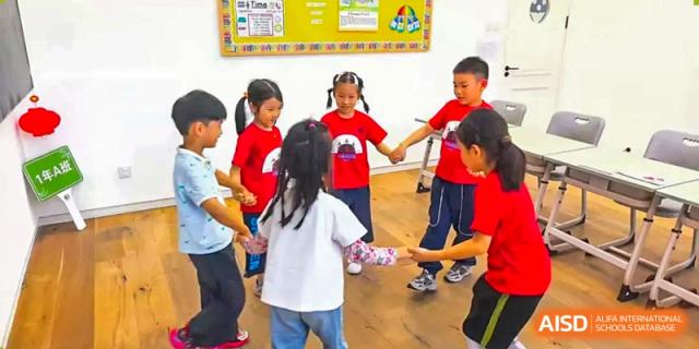 A group of young primary students in red and white shirts participate in a collaborative circle game on a wooden classroom floor at Beijing Tongzhou Shuren Private School.