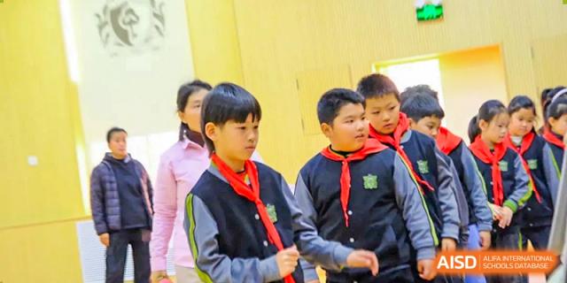 Students wearing black and grey school jackets with red scarves stand together during an indoor lesson, listening attentively to their instructor at Beijing Tongzhou Shuren Private School.