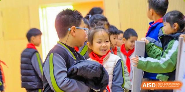 A young female student with a red scarf smiles brightly while standing among her classmates during an indoor school gathering at Beijing Tongzhou Shuren Private School.