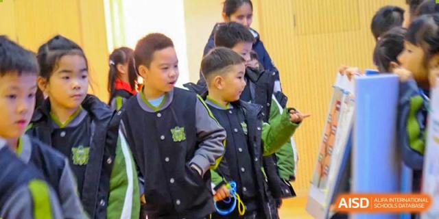 Two young boys in matching school jackets look toward an educational display board, pointing and discussing the information presented at Beijing Tongzhou Shuren Private School.