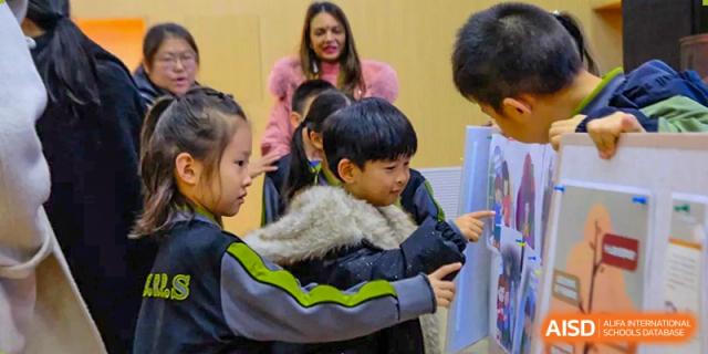Students gather around a teacher to look at a colorful educational poster, engaging in a hands-on learning moment inside the classroom at Beijing Tongzhou Shuren Private School.