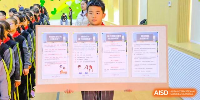 A student stands proudly while holding a large informational poster featuring various educational charts and diagrams for a class presentation at Beijing Tongzhou Shuren Private School.