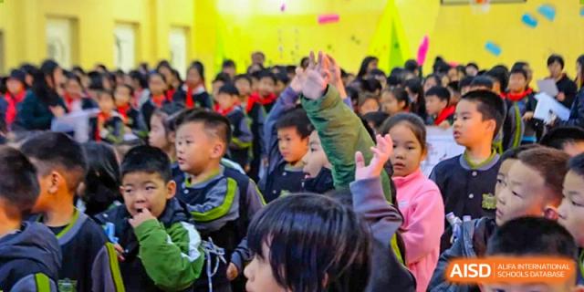 A large group of students gather in a brightly lit hall, with some raising their hands to participate in a lively school-wide Q&A session at Beijing Tongzhou Shuren Private School.