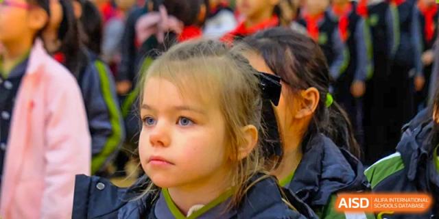 A close-up portrait of a young girl with blonde hair looking forward with a focused expression during a morning school assembly at Beijing Tongzhou Shuren Private School.