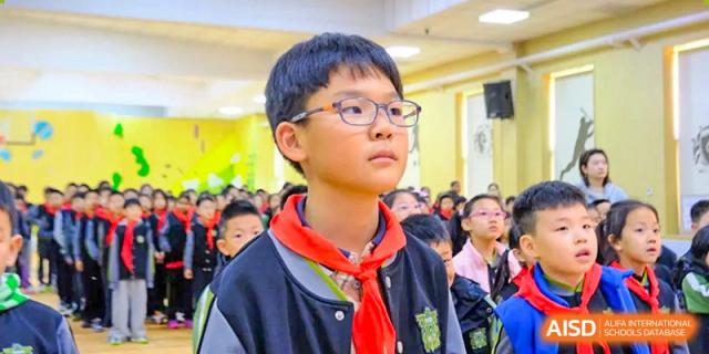 A focused young student with glasses and a red scarf stands attentively among his peers during a formal indoor assembly or commemorative event held at Beijing Tongzhou Shuren Private School.