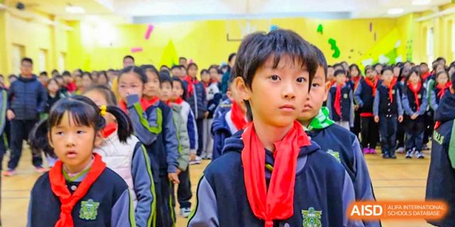 A young boy wearing a red scarf stands prominently in the foreground of a large group of students during an indoor school event at Beijing Tongzhou Shuren Private School.