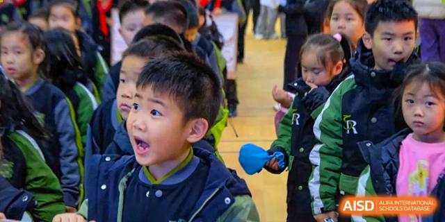 A young boy in a school uniform looks on with wide-eyed wonder and an expressive face during an engaging presentation or performance for the students at Beijing Tongzhou Shuren Private School.