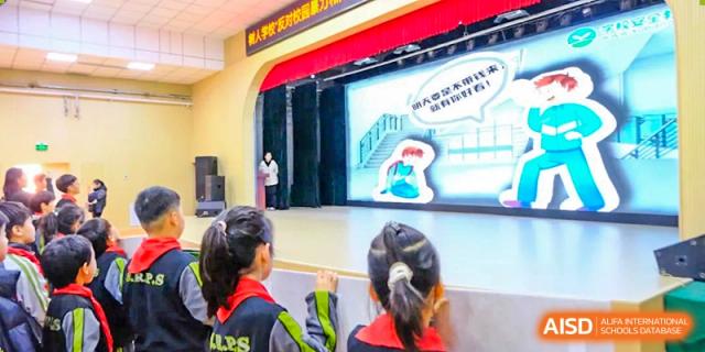 Students watch intently as a speaker presents on a large stage featuring a digital screen with illustrative educational graphics during a school gathering at Beijing Tongzhou Shuren Private School.
