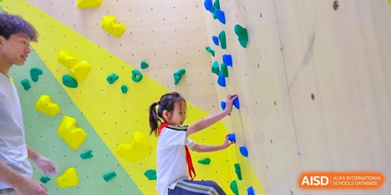 A young girl in a school uniform with a red scarf focuses intently as she climbs a bouldering wall, demonstrating strength and coordination at Beijing Tongzhou Shuren Private School.