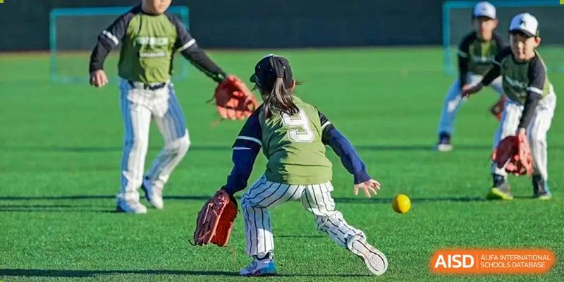 A student athlete in a green jersey and striped pants prepares to field a ball on the green turf during a competitive youth baseball match at Beijing Tongzhou Shuren Private School.