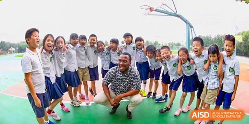 A coach sits on the ground surrounded by a large group of smiling students on an outdoor sports court, celebrating a day of fun and teamwork at Beijing Tongzhou Shuren Private School.