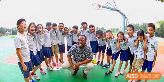 A coach sits on the ground surrounded by a large group of smiling students on an outdoor sports court, celebrating a day of fun and teamwork at Beijing Tongzhou Shuren Private School.