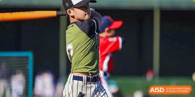 A young baseball batter in a green jersey and striped pants takes a powerful swing at the plate during an afternoon game on the school diamond at Beijing Tongzhou Shuren Private School.