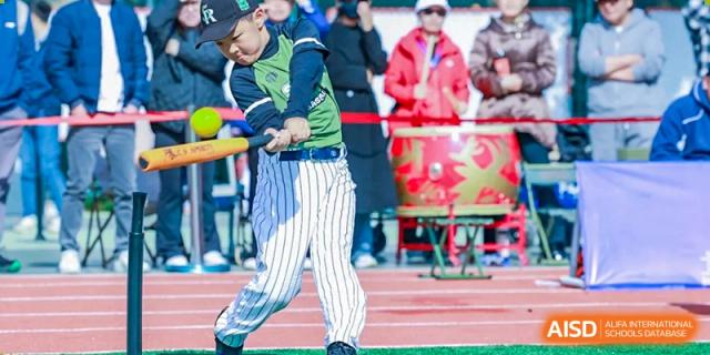 A young baseball player in a green and white striped uniform swings a yellow bat during an outdoor game while spectators watch from the sidelines at Beijing Tongzhou Shuren Private School.
