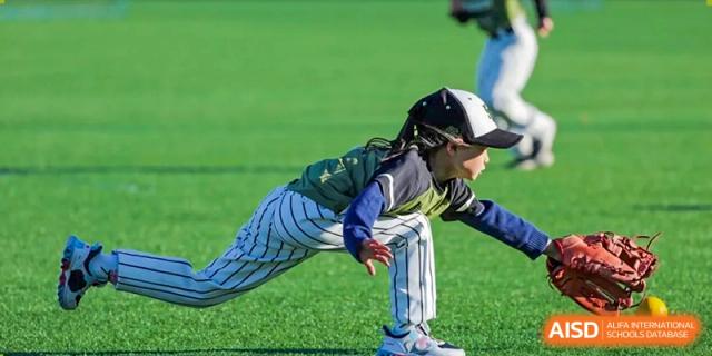 A young baseball player in a green jersey and striped pants lunges forward to make a catch during a high-energy outdoor sports competition at Beijing Tongzhou Shuren Private School.