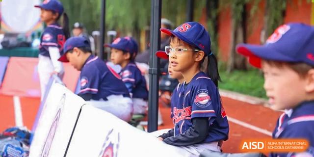 A young student athlete in a blue baseball cap and jersey sits in the dugout, watching the game intently and supporting his teammates at Beijing Tongzhou Shuren Private School.