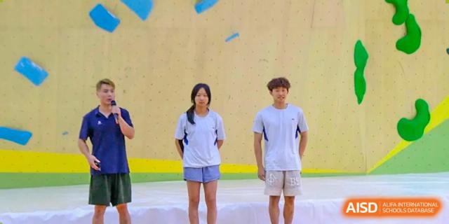 Three students stand on a stage in front of a climbing wall, with one speaking into a microphone during a school presentation or sports ceremony at Beijing Tongzhou Shuren Private School.
