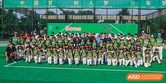 A large group of young baseball players in matching green uniforms poses together for a team photo on the field under a "Boom" banner at Beijing Tongzhou Shuren Private School.