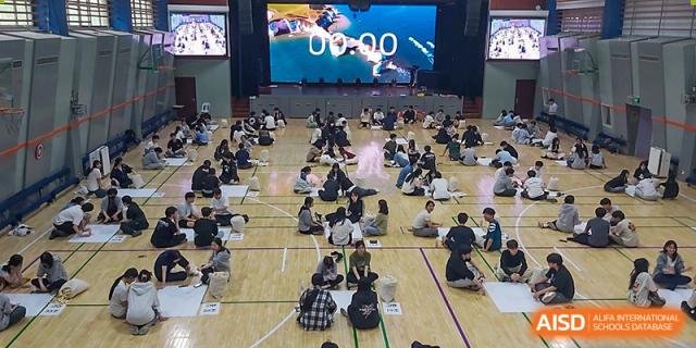 A high-angle view shows dozens of student teams spread across a large indoor gymnasium floor working together on creative wooden block projects at the Korean International School In Beijing.