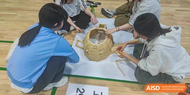 A group of students works together on the gym floor to assemble a rounded, hollow structure made of small wooden blocks during a collaborative design task at the Korean International School In Beijing.